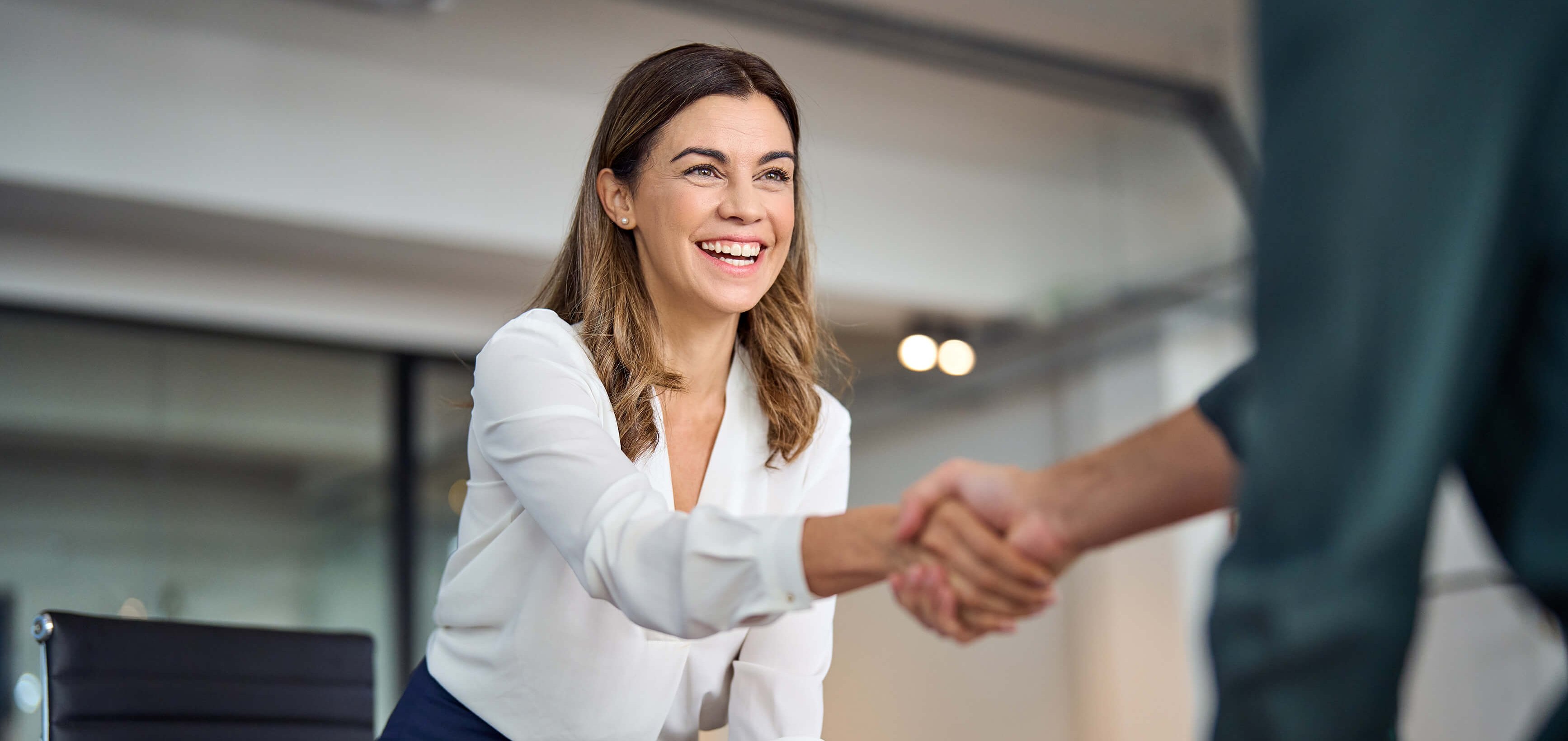 Mujer sonriente con blusa blanca estrecha la mano de otra persona en un entorno de oficina.