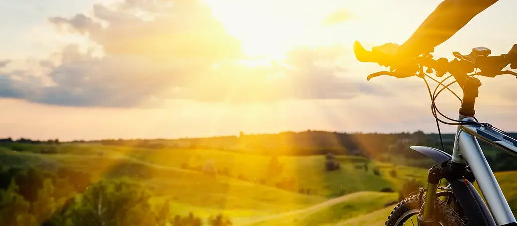 Bicicleta en primer plano durante un atardecer en el campo, con colinas verdes y rayos de sol iluminando el paisaje.