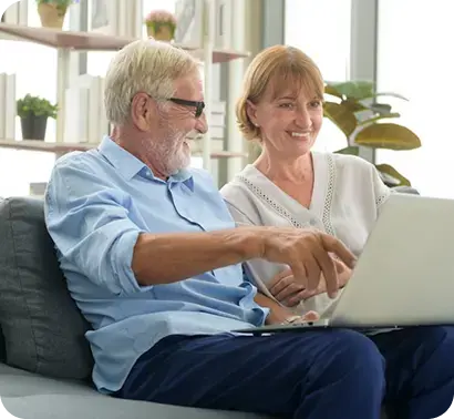 Pareja de adultos mayores sonriendo mientras utiliza una computadora portátil en el sofá.