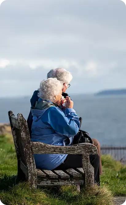 Pareja de adultos mayores sentada en un banco al aire libre contemplando el paisaje.