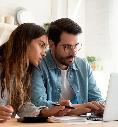 Pareja joven revisando información financiera en una laptop desde la cocina de su hogar, con calculadora y bolígrafo sobre la mesa.