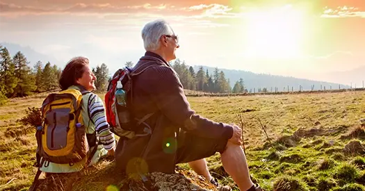 Pareja de adultos mayores con mochilas sentados al aire libre contemplando el paisaje al atardecer