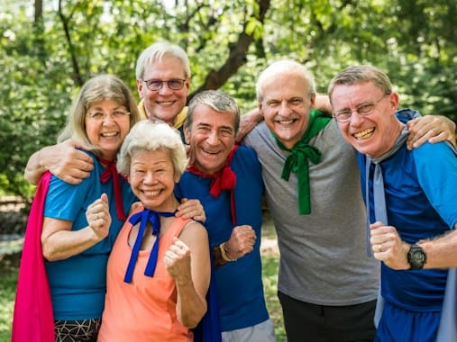 Grupo de adultos mayores sonrientes con capas de superhéroes posando juntos y celebrando con los puños en alto en un parque al aire libre.