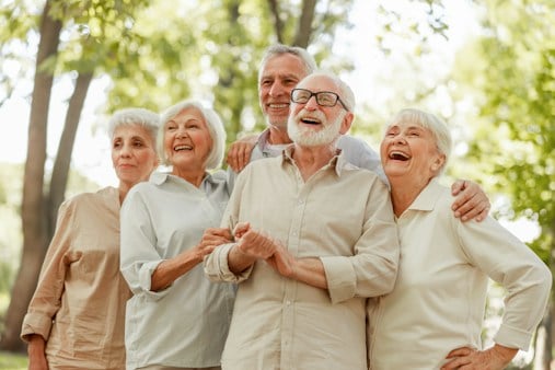 Grupo de cinco adultos mayores sonriendo y abrazados al aire libre en un parque, vistiendo ropa casual en tonos claros con árboles de fondo.