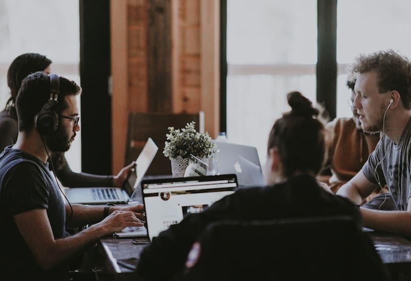 Grupo de personas reunidas alrededor de una mesa trabajando en computadoras portátiles y usando audífonos en un espacio de coworking.