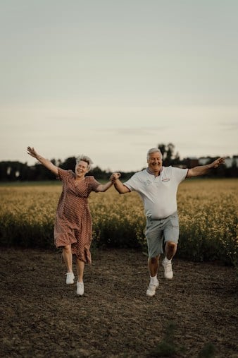 Pareja de adultos mayores tomados de la mano mientras corren y saltan sonrientes en un campo abierto con flores amarillas de fondo.