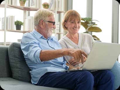 Pareja de adultos mayores sentada en un sofá, sonriendo mientras utiliza una computadora portátil.