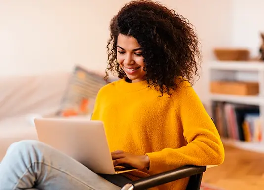 Mujer sonriente con suéter amarillo utiliza una laptop mientras se sienta en una sala acogedora.