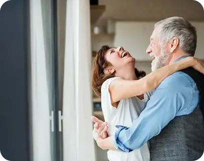 Hombre adulto mayor y mujer sonriendo mientras se abrazan junto a una ventana.