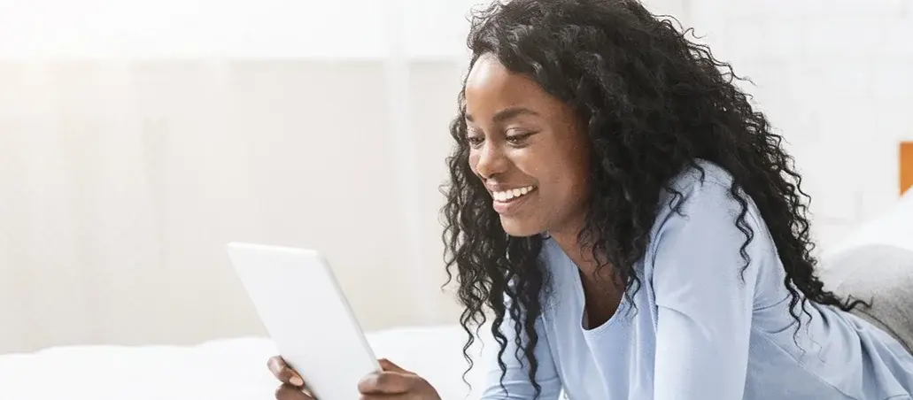 Mujer sonriente usando una tableta mientras está recostada en la cama.