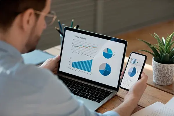 Hombre revisando gráficos estadísticos en una laptop y un teléfono móvil desde su escritorio, con enfoque en análisis financiero.